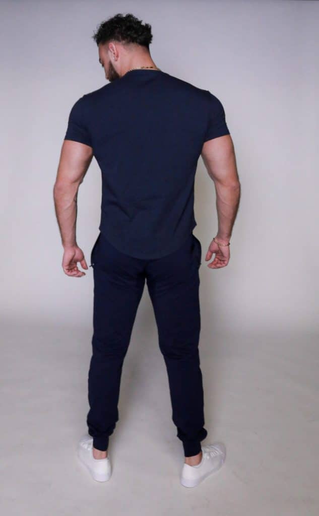 A man with short dark hair stands facing away from the camera. He is wearing a fitted dark blue t-shirt, dark blue pants, and white sneakers, against a plain light background.
