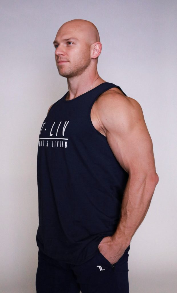 A muscular, bald man with fair skin wearing a navy blue sleeveless shirt and matching pants stands with one hand in his pocket, facing slightly to the left against a plain background.