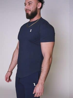 A man with a beard, wearing a navy blue fitted t-shirt and matching joggers, stands facing slightly to the left against a plain light background.