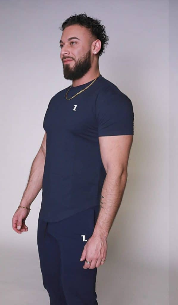 A man with a beard, wearing a navy blue fitted t-shirt and matching joggers, stands facing slightly to the left against a plain light background.