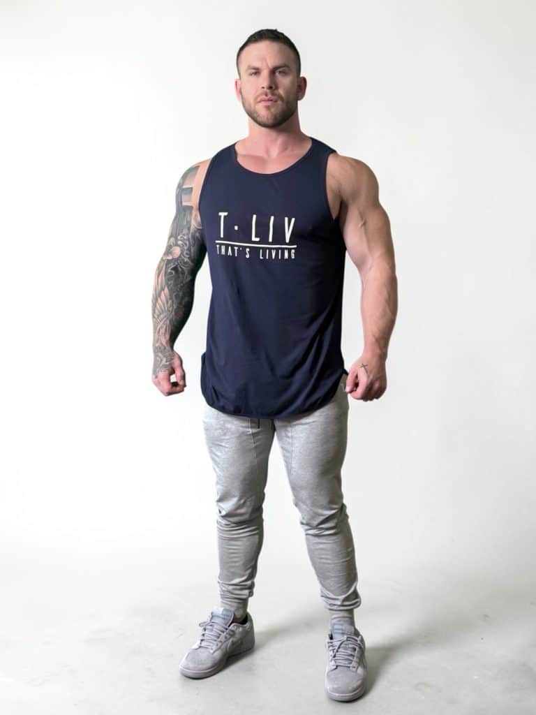 A muscular man with tattoos on his left arm stands against a plain white background, wearing a navy blue tank top with "T-LIV THAT'S LIVING" on it, gray joggers, and gray sneakers. He looks directly at the camera.
