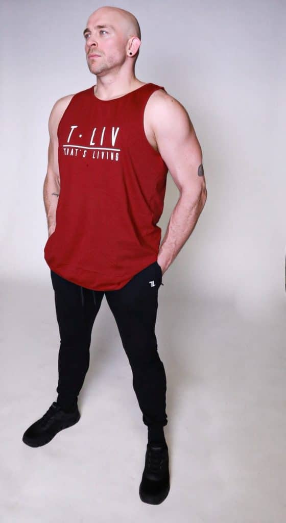 A bald man wearing a sleeveless red shirt and black pants stands with his hands in his pockets, looking slightly upward against a plain, light background.