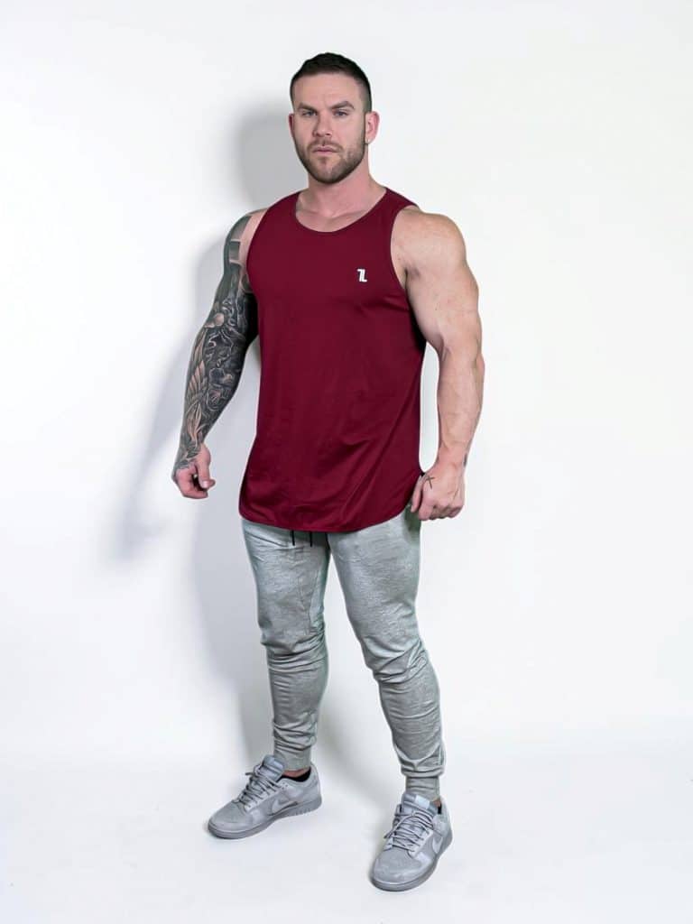 A muscular man with a tattooed right arm wears a sleeveless maroon tank top, light gray jogger pants, and gray sneakers, standing against a plain white background.
