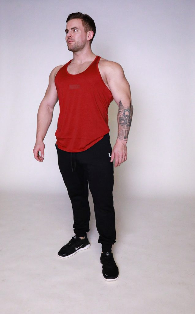 A muscular man wearing a red sleeveless tank top, black joggers, and black sneakers stands against a plain light background, looking slightly to his left with his arms relaxed at his sides.