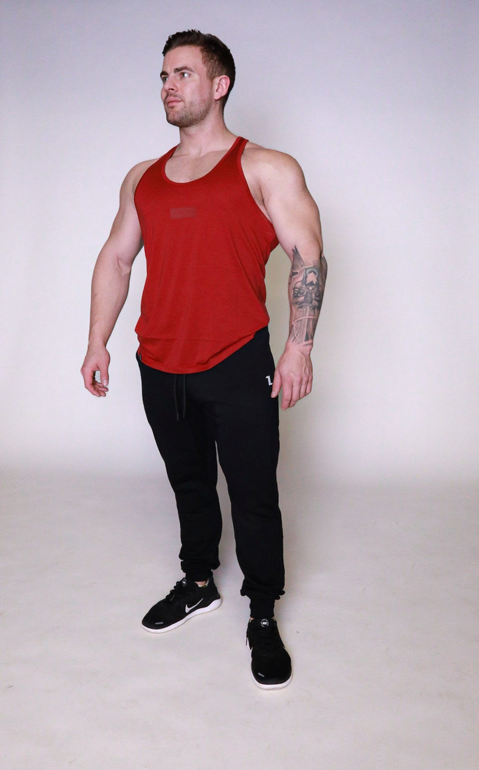 A muscular man wearing a red sleeveless tank top, black joggers, and black sneakers stands against a plain light background, looking slightly to his left with his arms relaxed at his sides.