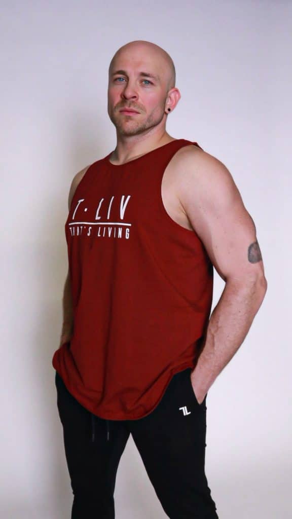 A muscular bald man with a trimmed beard stands confidently, wearing a sleeveless maroon shirt and black pants. His hands are in his pockets and he has a neutral expression, posing against a plain light background.