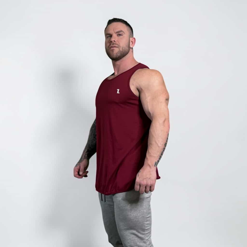 A muscular man wearing a maroon sleeveless tank top and light gray sweatpants stands against a plain white background, looking confidently at the camera.
