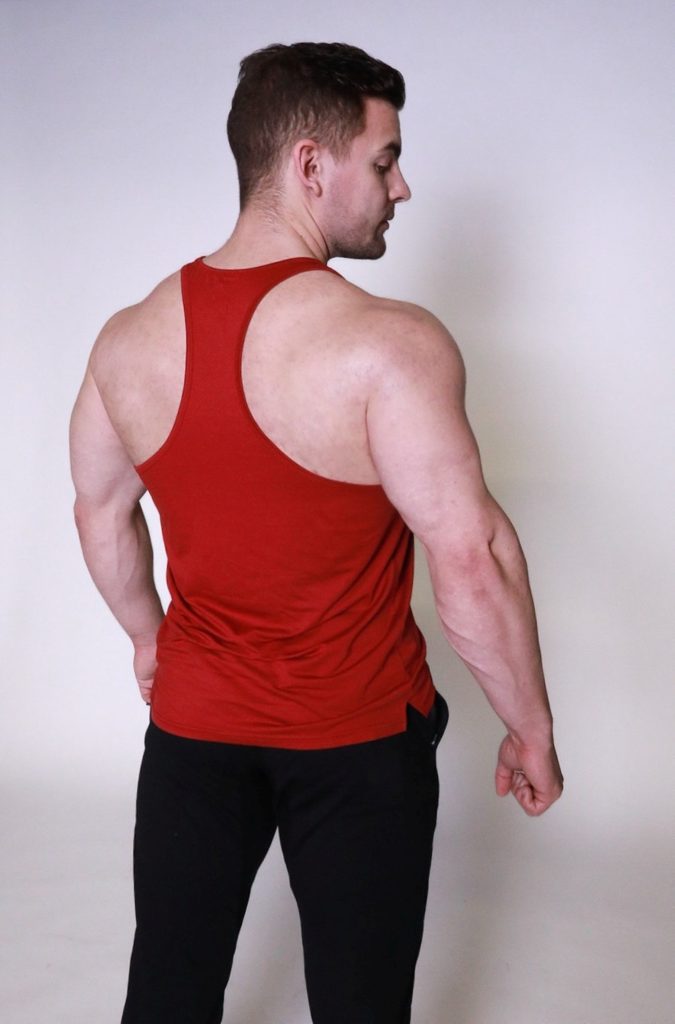 A muscular man in a red racerback tank top and black pants stands facing away, showing his back and shoulder muscles against a plain light background.