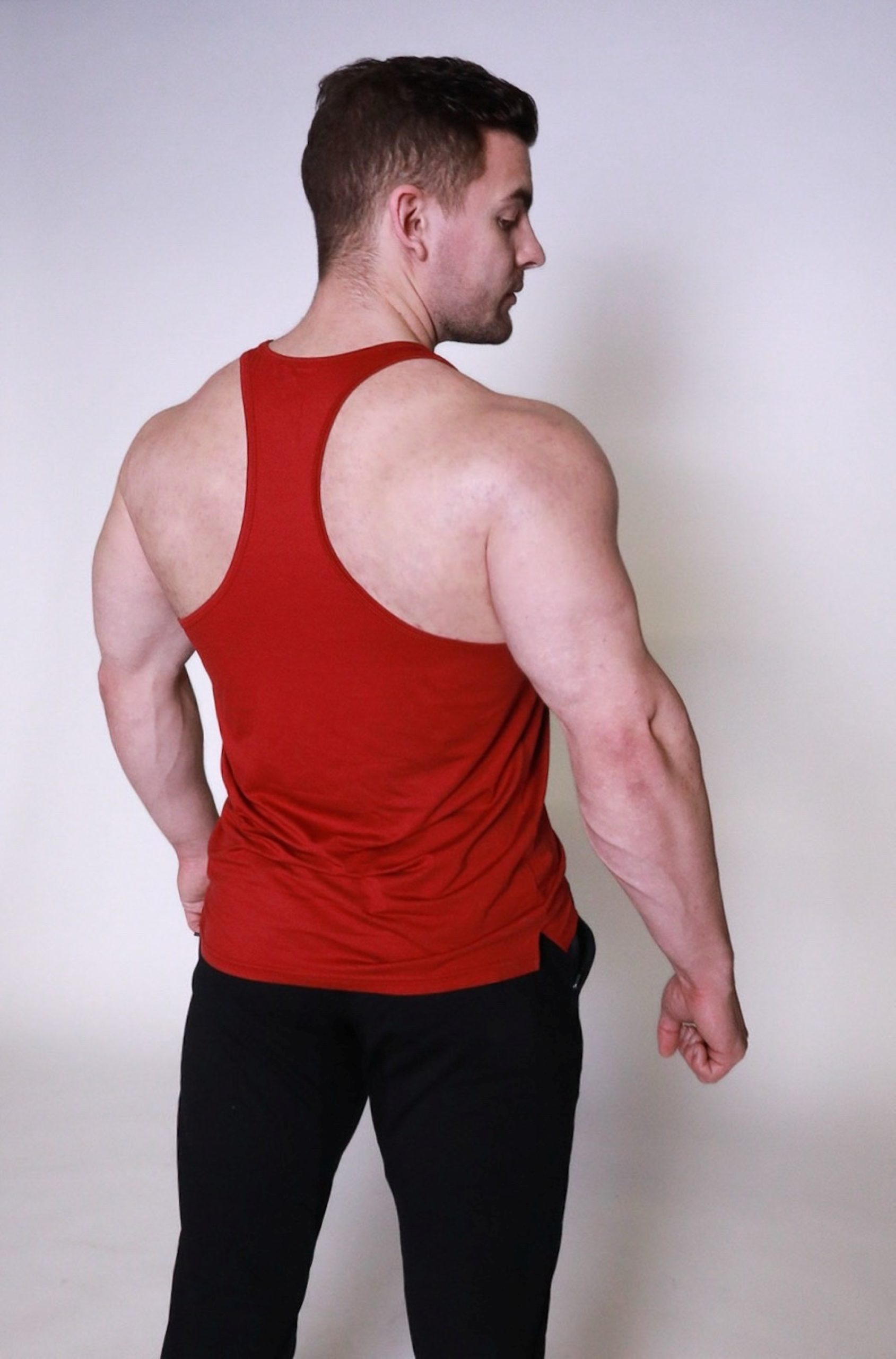 A muscular man in a red racerback tank top and black pants stands facing away, showing his back and shoulder muscles against a plain light background.