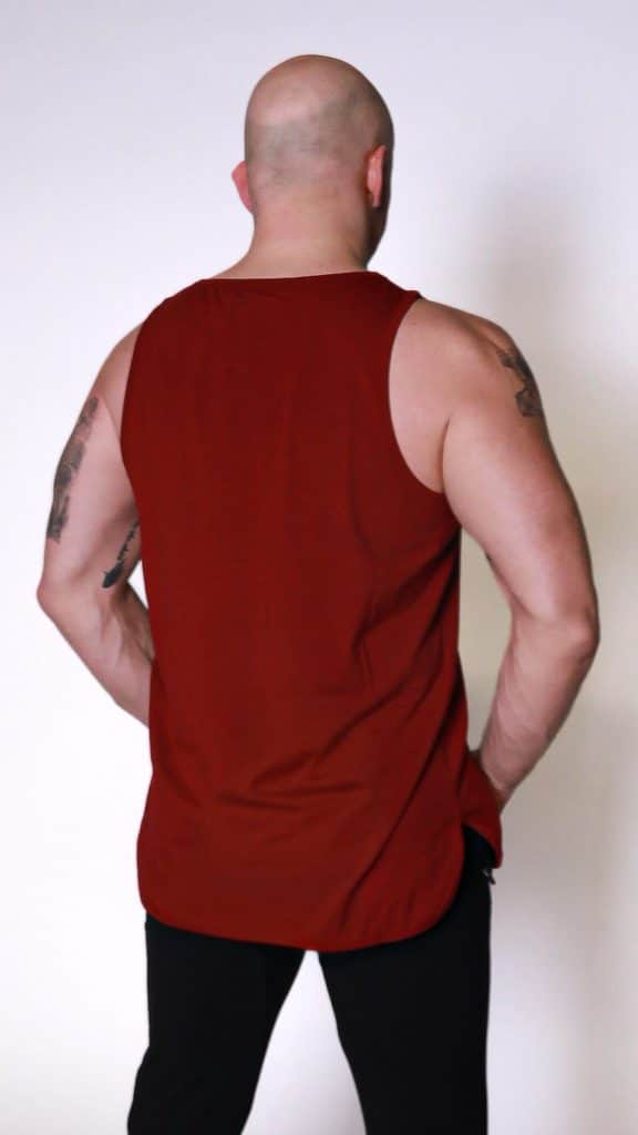 A bald man with tattoos on his arms is standing with his back to the camera, wearing a sleeveless dark red tank top and black pants against a plain light background.