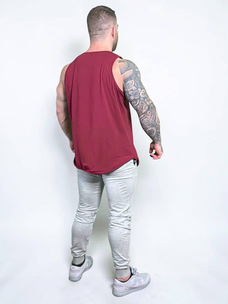 A muscular person stands facing away, wearing a sleeveless maroon shirt, light gray jogger pants, and gray sneakers. Their right arm has a detailed tattoo sleeve. The background is plain white.