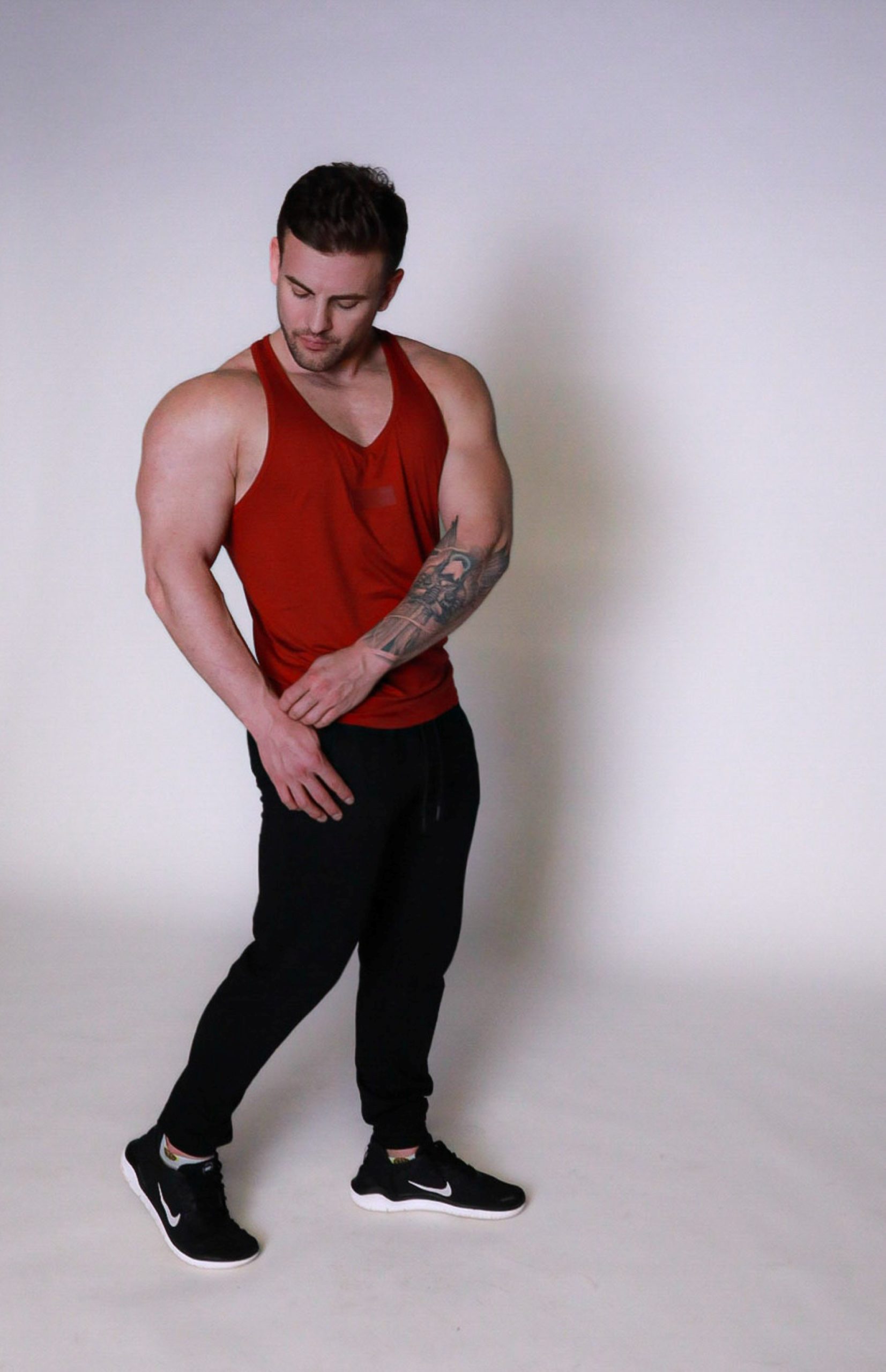 A muscular man in a red tank top, black joggers, and black sneakers stands against a plain white background, looking down and to his left with one hand on his thigh and one hand near his waist.