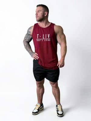 A muscular man with tattoos wears a red sleeveless shirt that says "T. LIV THAT'S LIVING," black shorts, and sneakers, standing on a white background and looking off to the side.