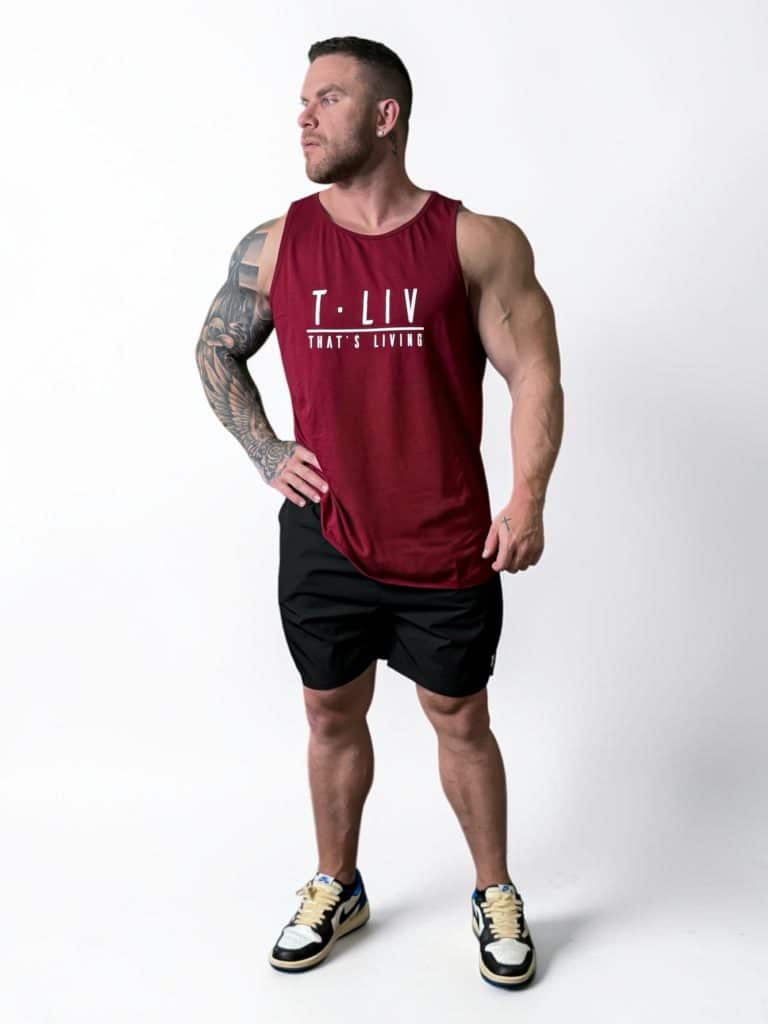 A muscular man with tattoos wears a red sleeveless shirt that says "T. LIV THAT'S LIVING," black shorts, and sneakers, standing on a white background and looking off to the side.
