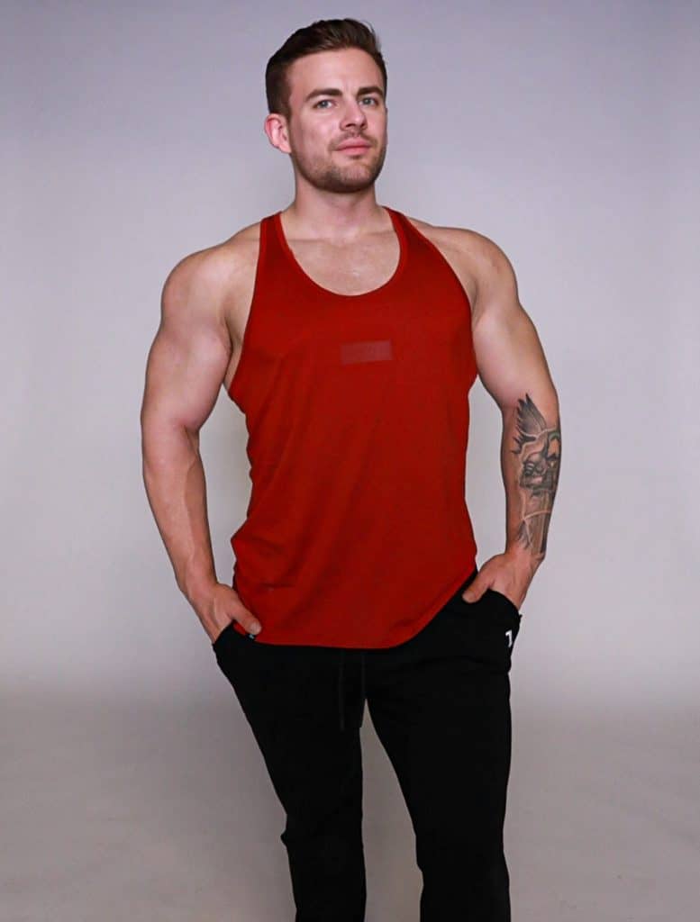 A muscular man wearing a red sleeveless tank top and black pants stands with his hands in his pockets against a plain light background. He has short hair and a tattoo on his left forearm.
