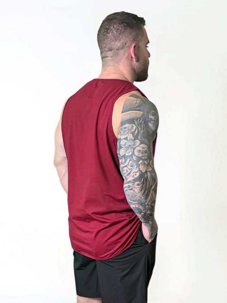 A man with short hair, wearing a sleeveless maroon shirt and black shorts, stands facing away. His right arm is fully covered with a detailed floral tattoo. The background is plain and white.