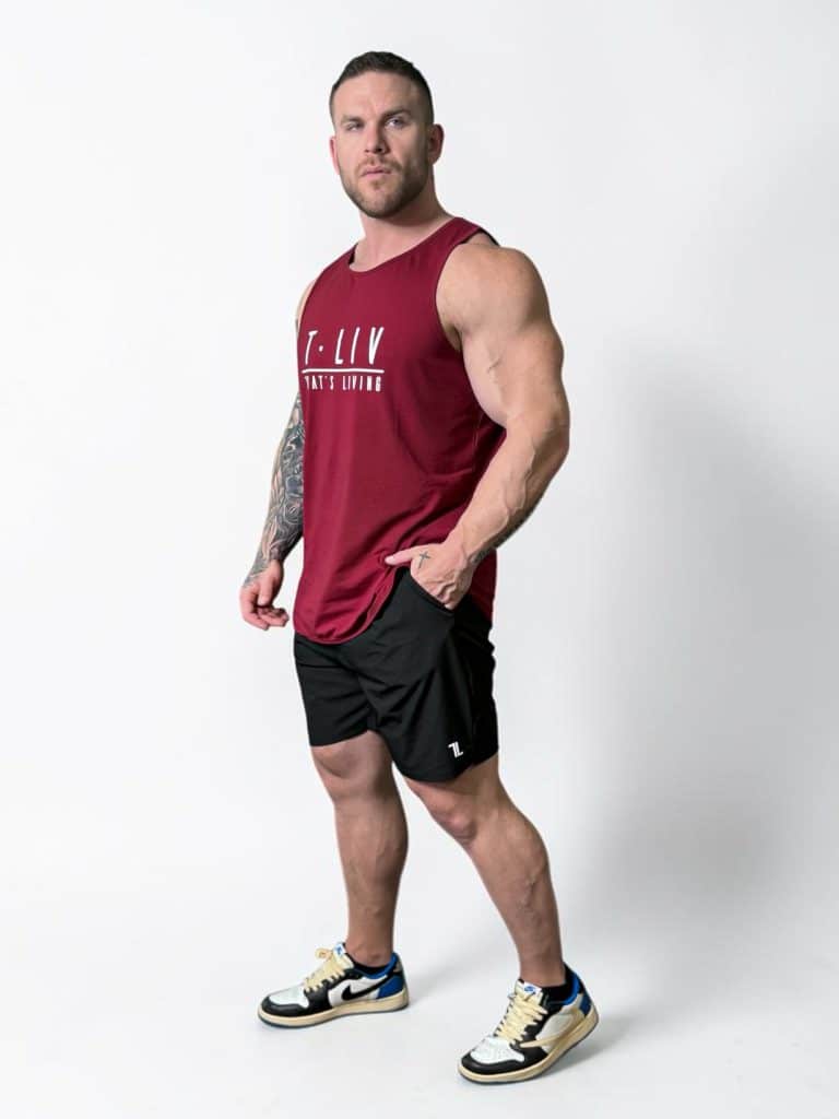A muscular man stands against a plain white background wearing a sleeveless maroon shirt, black athletic shorts, and white sneakers with black and gold accents. He has a tattooed right arm and a serious expression.