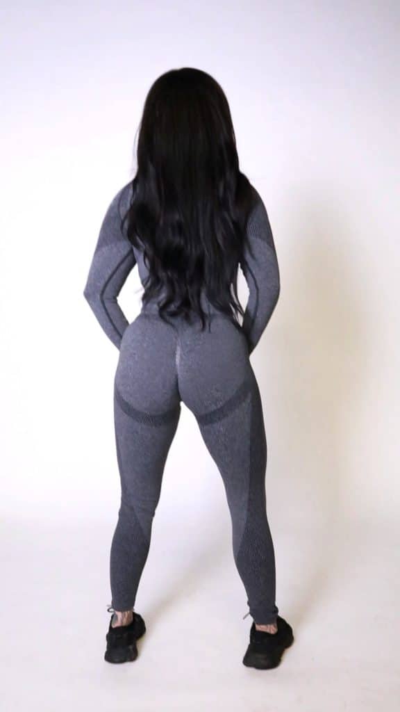 A person with long dark hair, dressed in a fitted gray athletic outfit and black sneakers, stands facing away against a plain white background.