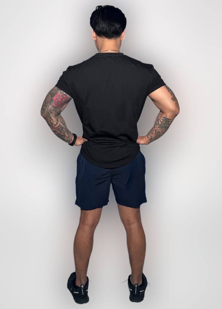 A man with short dark hair stands facing away, hands on hips, wearing a black t-shirt, navy shorts, black sneakers, and displaying tattoos on both arms, against a plain white background.