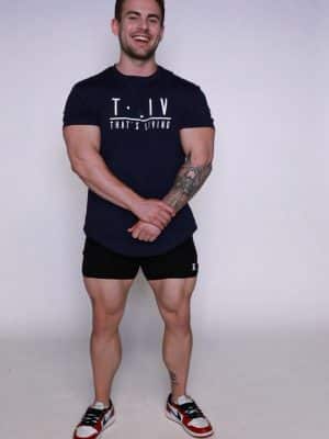 A smiling man with short brown hair stands against a plain light background, wearing a navy T-shirt with "T.LIV THAT'S LIVING" text, black shorts, red and white sneakers, and showing a tattoo on his left forearm.