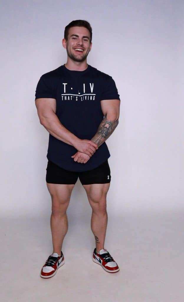 A smiling man with short brown hair stands against a plain light background, wearing a navy T-shirt with "T.LIV THAT'S LIVING" text, black shorts, red and white sneakers, and showing a tattoo on his left forearm.