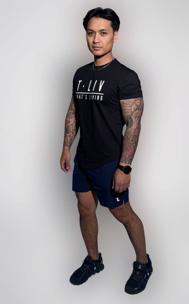 A man with tattooed arms stands in a studio wearing a black T-shirt with "T-LIV WHAT'S LIVING" printed on it, navy shorts, black sneakers, and a watch, smiling slightly and facing towards the camera.