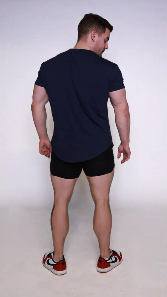 A man stands facing away from the camera, wearing a navy t-shirt, black shorts, and red-and-white sneakers, against a plain white background.