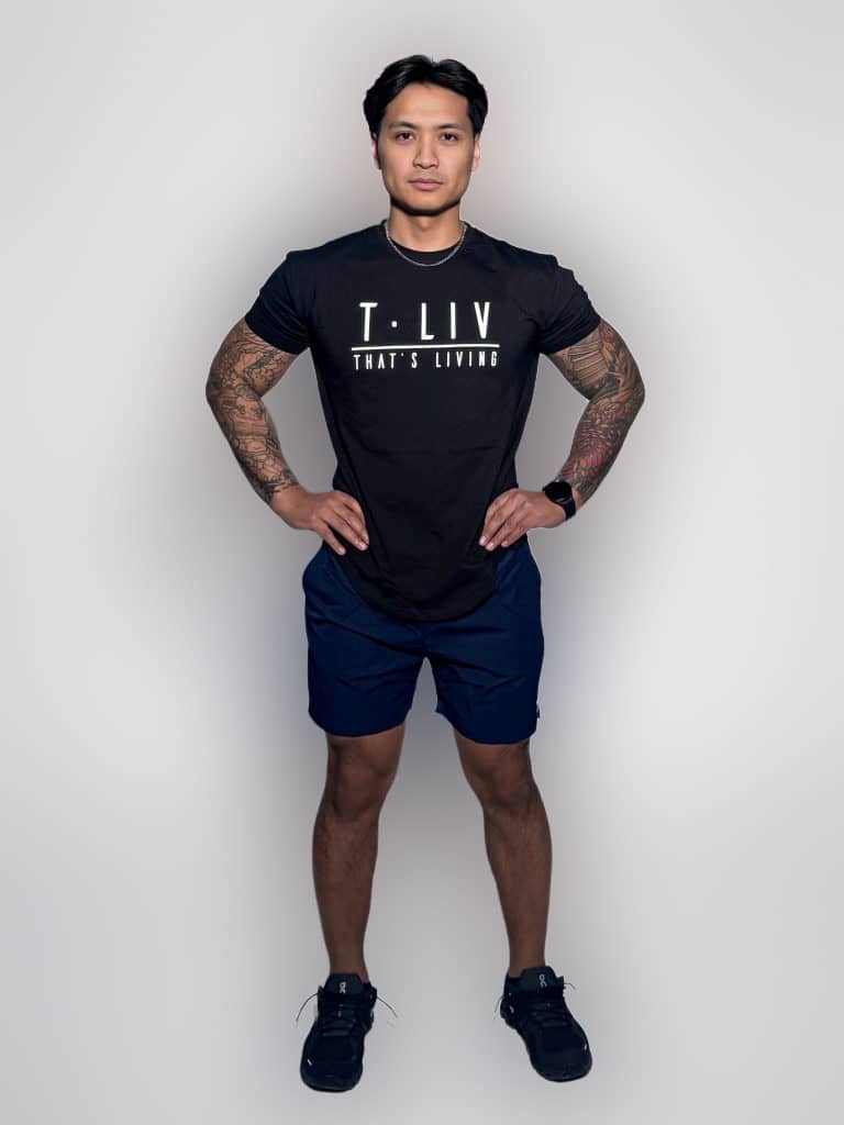 A man stands confidently with hands on hips, wearing a black "T-LIV THAT'S LIVING" t-shirt, navy shorts, and black sneakers, against a plain light background.