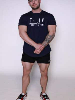 A muscular man stands against a plain white background, wearing a navy "T.LIV THAT'S LIVING" t-shirt, black shorts, and red, white, and blue sneakers. He has tattoos on his left forearm and lower right leg.