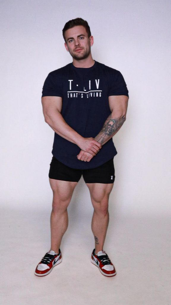 A muscular man stands against a plain white background, wearing a navy "T.LIV THAT'S LIVING" t-shirt, black shorts, and red, white, and blue sneakers. He has tattoos on his left forearm and lower right leg.