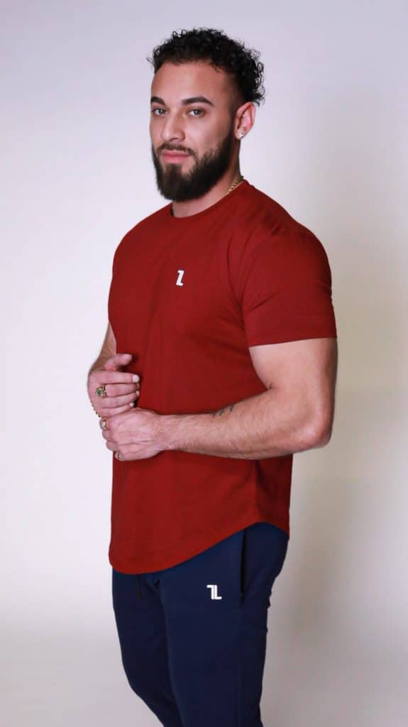 A man with a beard and curly hair wears a red T-shirt and navy blue pants, both featuring a small white "L" logo, standing against a plain light background.