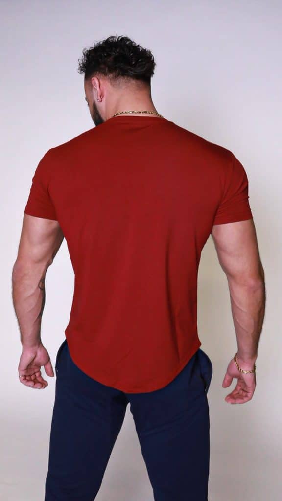 A muscular man with short, dark curly hair is standing with his back to the camera, wearing a fitted red t-shirt, dark pants, and a gold chain necklace and bracelet, against a plain light background.