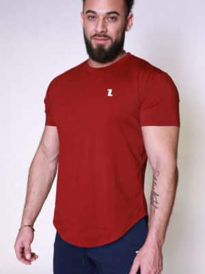 A man with a beard and curly hair is standing against a plain light background, wearing a fitted red T-shirt with a small white logo and dark pants. He is looking at the camera with a neutral expression.