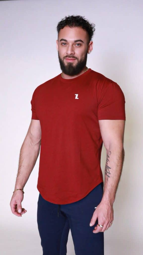 A man with a beard and curly hair is standing against a plain light background, wearing a fitted red T-shirt with a small white logo and dark pants. He is looking at the camera with a neutral expression.