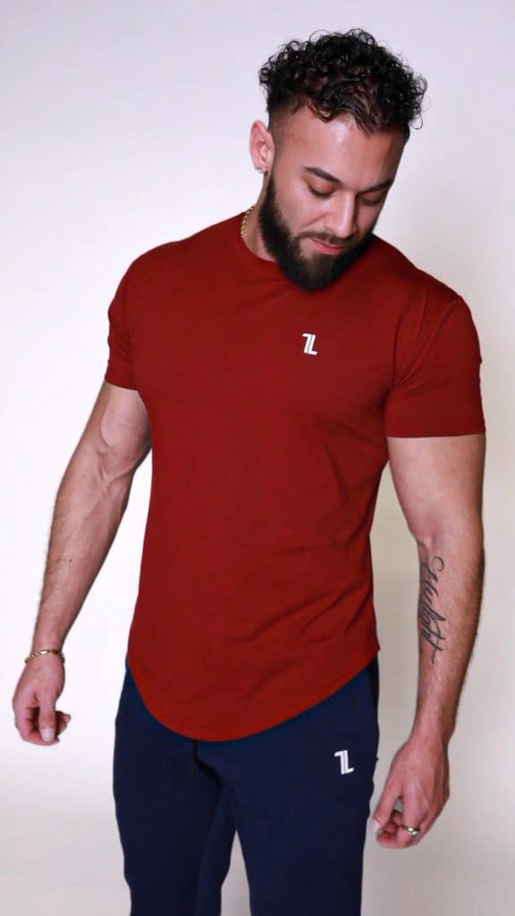 A man with a beard and curly hair stands against a plain white background, wearing a fitted red T-shirt and dark blue pants, both with a small white logo. He is looking down and slightly to his left, with his arms relaxed at his sides.